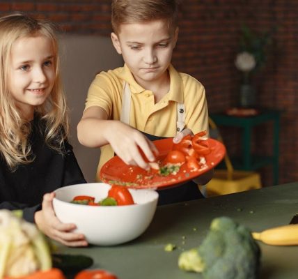 Positive little girl and boy mixing cutted ingredients for salad in big bowl while cooking at table with assorted fresh vegetables and fruits
