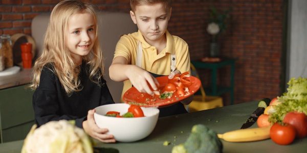 Positive little girl and boy mixing cutted ingredients for salad in big bowl while cooking at table with assorted fresh vegetables and fruits